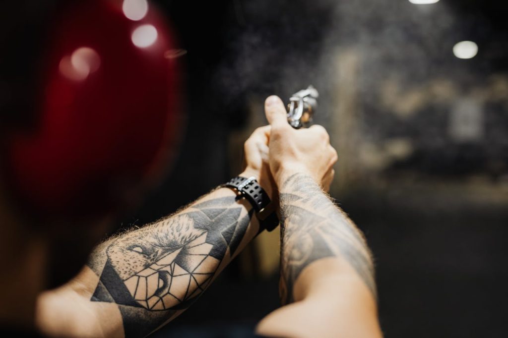 Close-up of tattooed arms aiming a gun at an indoor firing range with visible smoke.